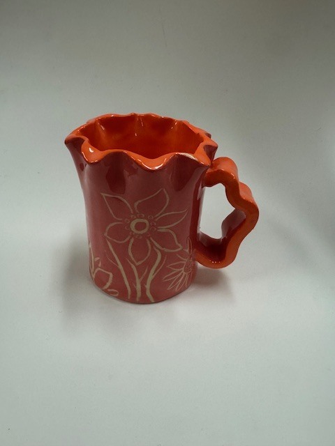 A small handmade ceramic mug with a glossy reddish‑orange glaze, a wavy rim, and a sculpted handle. The side features an etched flower design in a lighter clay color.