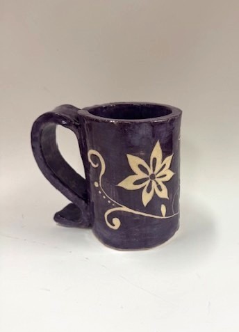 A dark purple ceramic mug with a large handle and a cream‑colored floral design carved or painted onto the side.