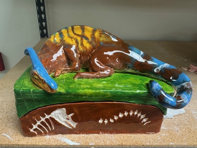 A ceramic sculpture of a lizard‑like creature lying on a green surface atop a rectangular base. The animal has a brown body with white and yellow markings and a curled blue tail. The base is decorated with white fossil‑style designs.