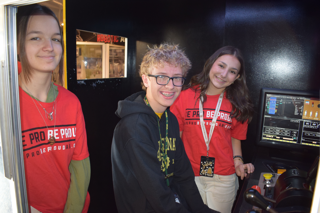 Three students stand inside a dark booth with a computer monitor displaying a robotics simulation interface. Two wear red “Be Pro Be Proud” shirts.