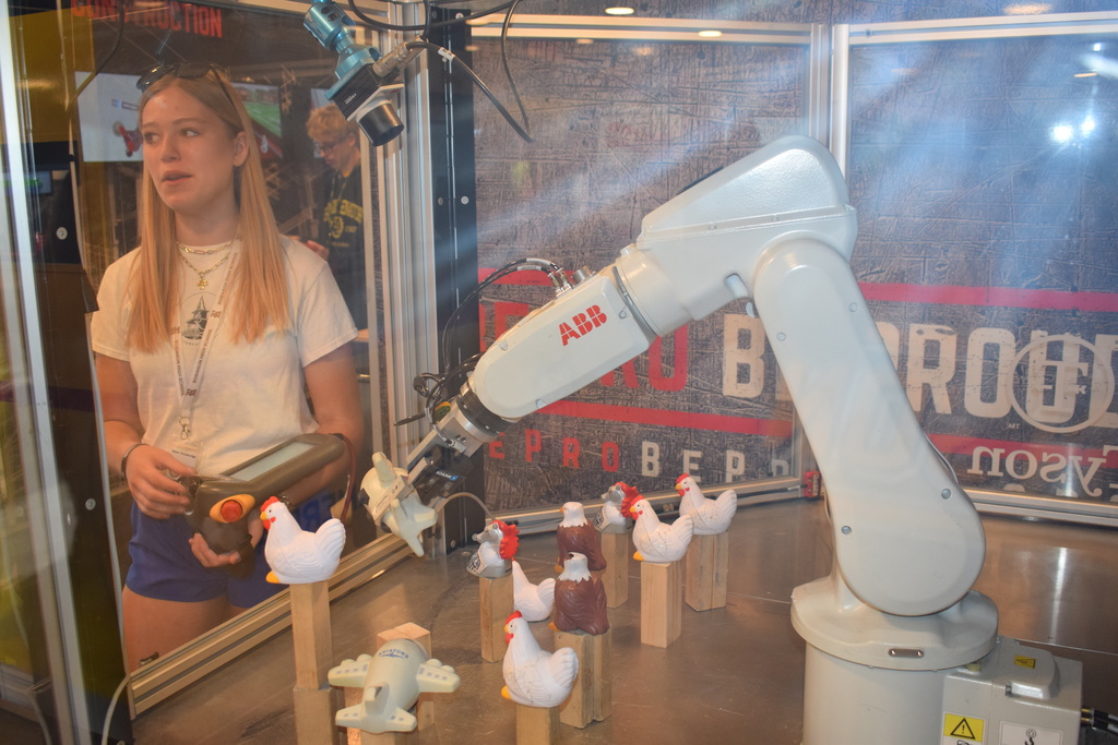 A student stands beside an ABB robotic arm inside a clear safety enclosure, holding a handheld controller. Small toy chickens and wooden blocks sit on the table inside the enclosure.