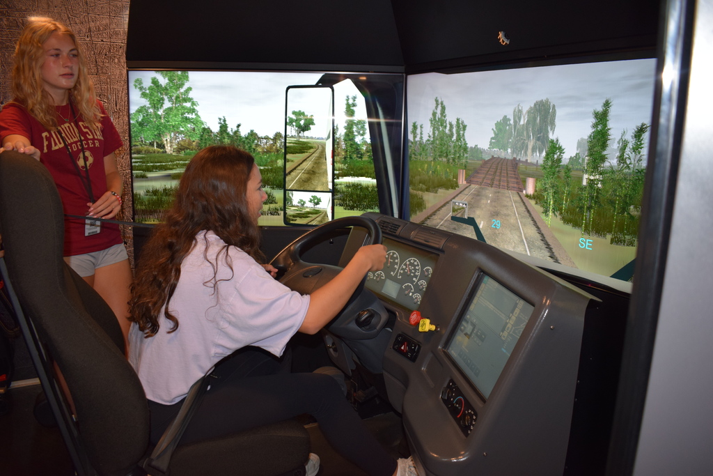 A student sits in a truck‑driving simulator holding the steering wheel while another student stands beside them. A large screen displays a road and bridge in the simulated environment.