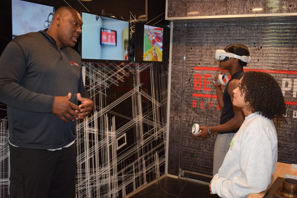 A student wearing a VR headset holds two controllers while another student observes. Two monitors behind them display simulation graphics, and the wall reads “Be Pro Be Proud.”