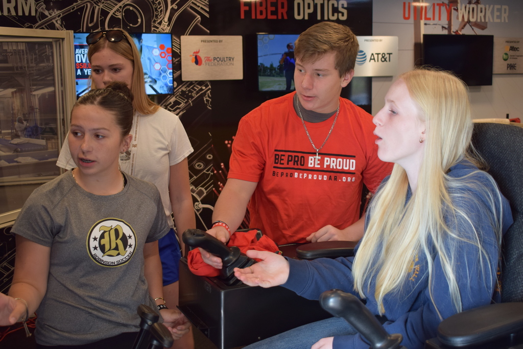Four students interact with a hands‑on simulator setup. Two are seated using joystick controls while two stand nearby. Signs behind them read “Fiber Optics,” “Utility Worker,” and “Be Pro Be Proud.”