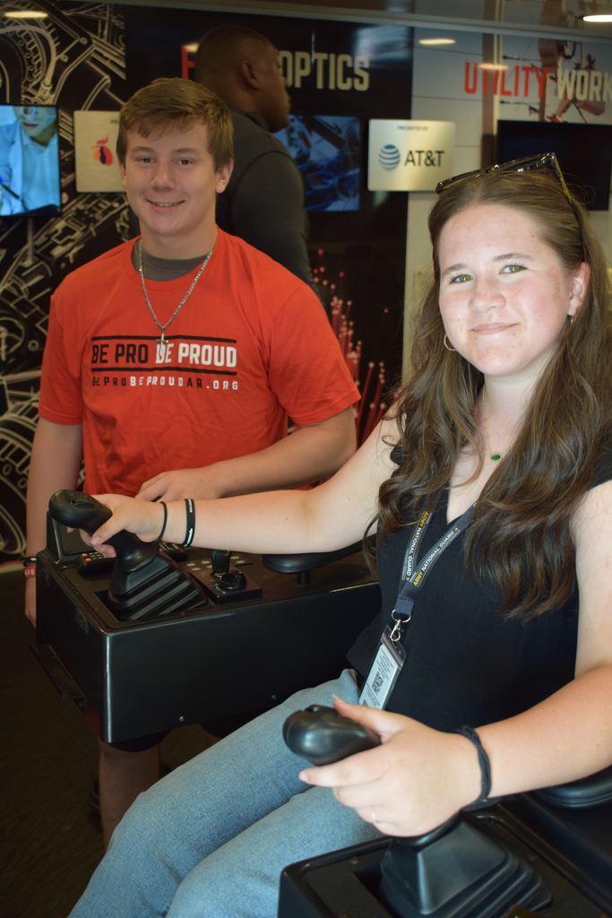 Two students operate joystick‑based simulators. One wears an Army National Guard lanyard, and the other wears a red “Be Pro Be Proud” shirt. Technical trade‑related signage is visible behind them.