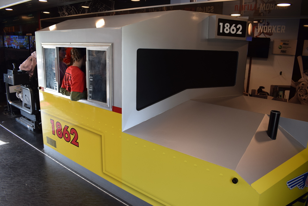 A large yellow train‑cab simulator labeled “1862” sits indoors with people visible inside the cab. Screens and equipment surround the exhibit.