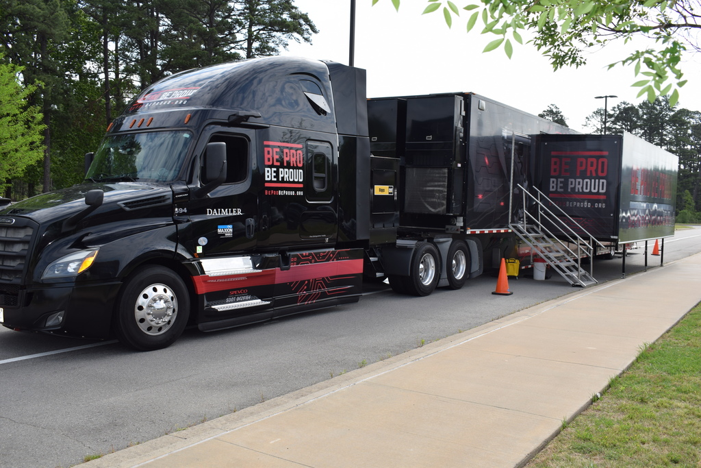 A black semi‑truck and trailer branded “Be Pro Be Proud” is parked outdoors with stairs leading into the trailer. Orange cones mark the entrance area.