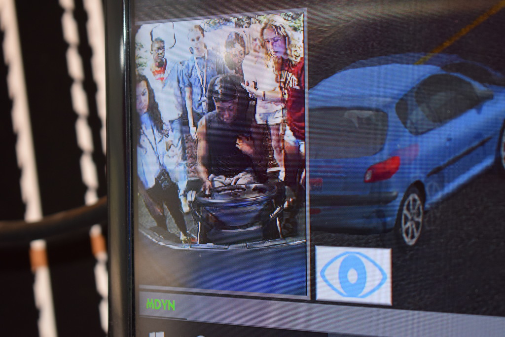 A screen shows two camera views: a group of people gathered outdoors on the left and a blue car on a roadway on the right. A blue eye icon and the text “HDYN” appear at the bottom.