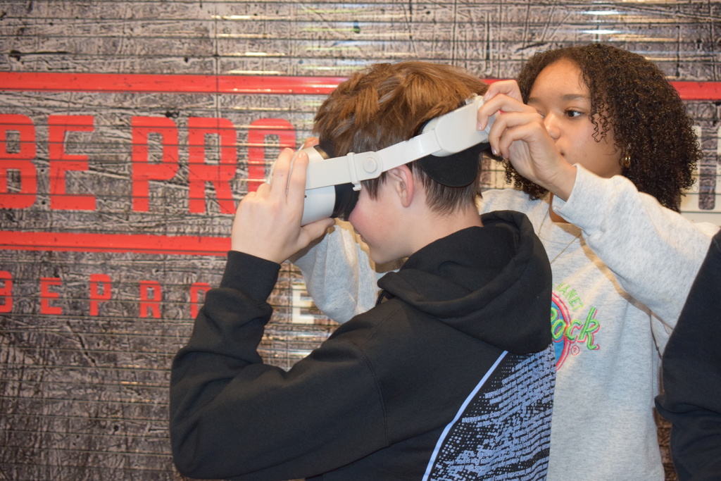 Two students stand in front of a wall labeled “Be Pro.” One student adjusts a white VR headset on the other.