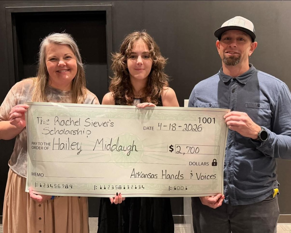 A Sylvan Hills High School senior, Hailey Middaugh, stands between two adults while holding a large ceremonial check for the Rachel Sievers Scholarship. All three are smiling. The check lists the award amount of $2,700 and is issued by Arkansas Hands & Voices.
