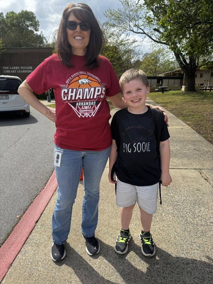 An adult and a child stand outside a library building. The adult wears a red Arkansas basketball shirt; the child wears a black “Pig Sooie” shirt.