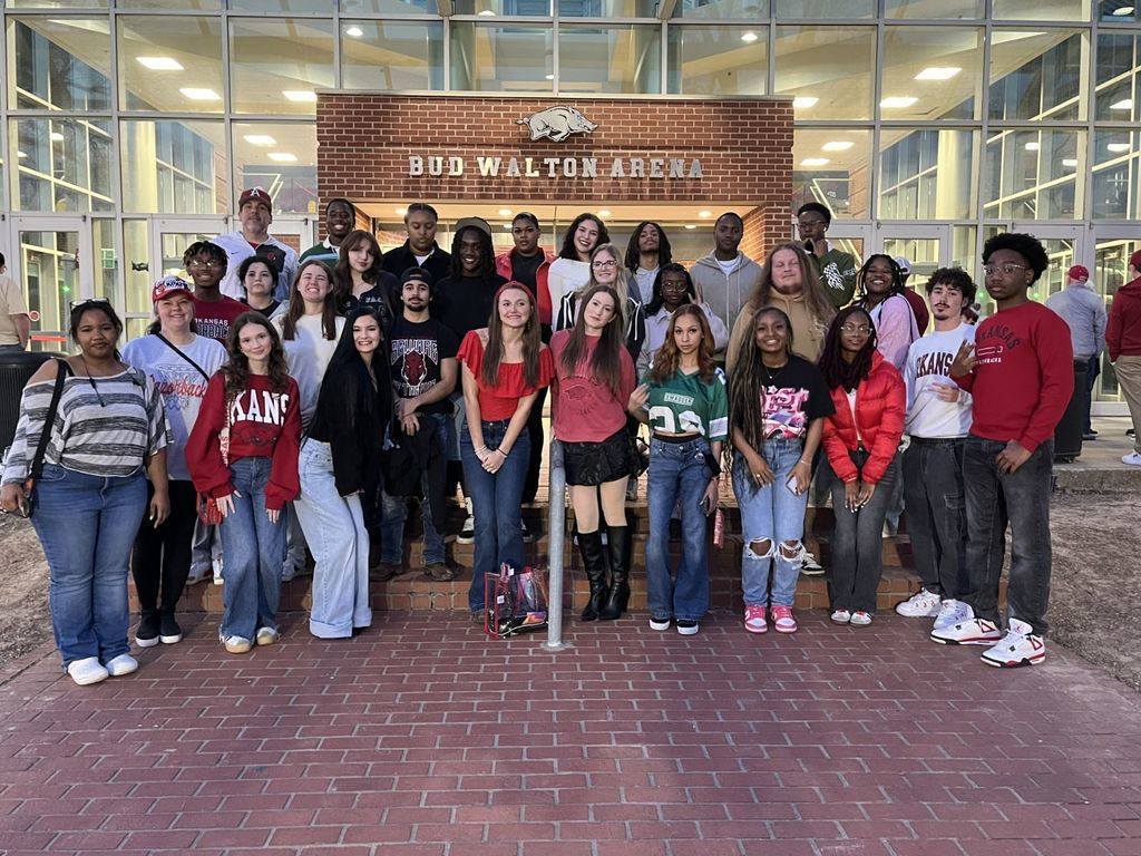 A group of people stand outside Bud Walton Arena wearing Arkansas Razorbacks apparel, posing for a photo.