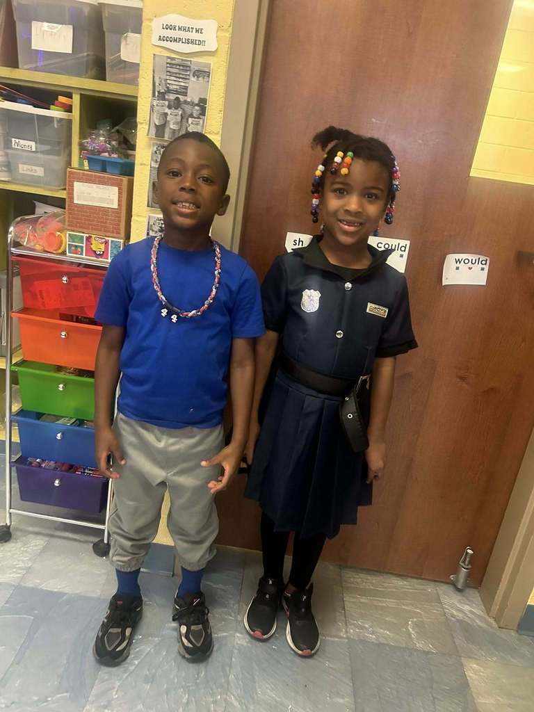Two students stand in a classroom near a door labeled with the words “should,” “could,” and “would.” One student wears a blue shirt and beaded necklace; the other wears a navy uniform dress with colorful hair beads.