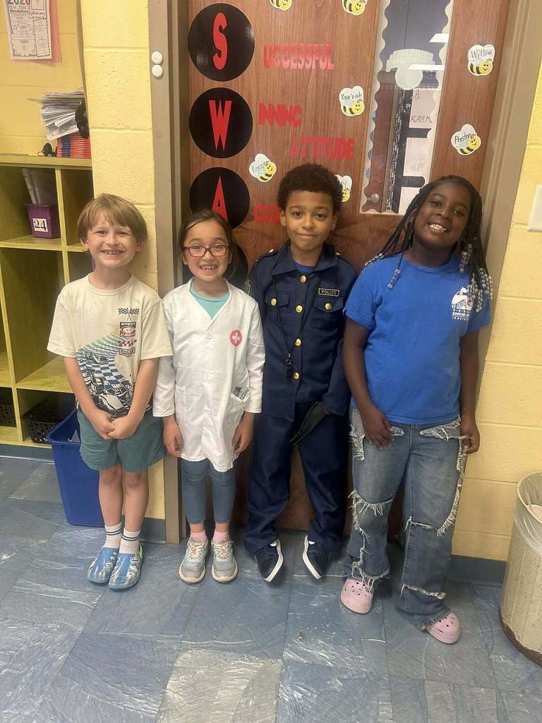 Four elementary students stand in front of a classroom door decorated with the phrase “Successful Winning Attitude.” Each child is dressed to represent a different future career, including a doctor and a police officer.