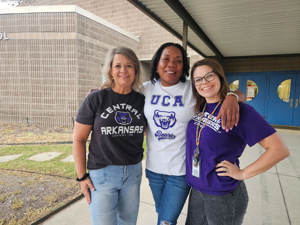 Three adults stand outside a school building wearing University of Central Arkansas shirts, smiling and showing college pride.