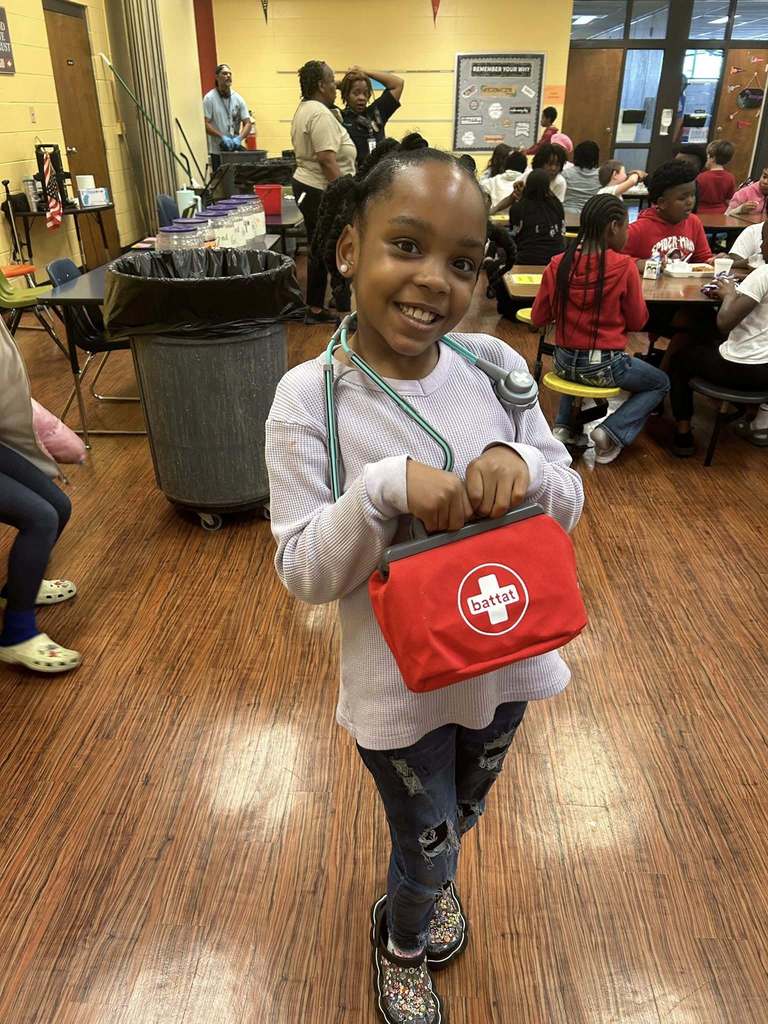 A student stands in a cafeteria wearing a toy stethoscope and holding a red toy medical bag, dressed as a future healthcare professional.
