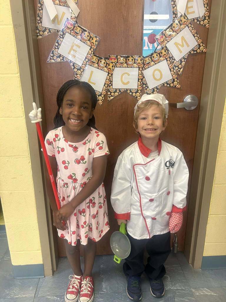 Two students stand in front of a classroom door with a “WELCOME” banner. One wears a pink dress with a cherry pattern; the other is dressed as a chef with a white jacket, chef hat, and small frying pan.