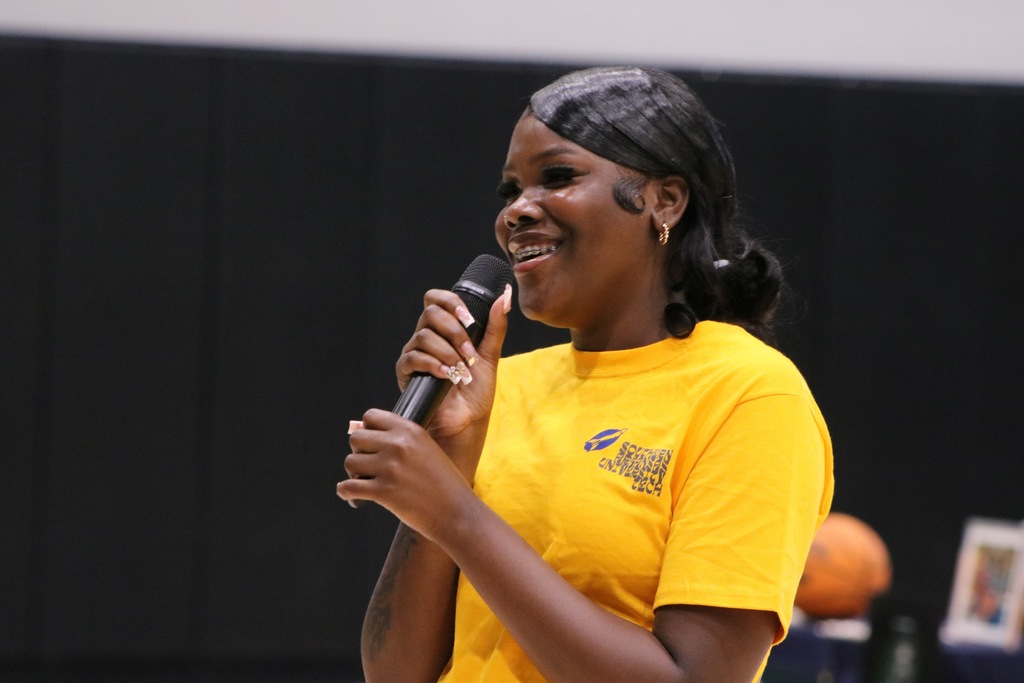 A student wearing a bright yellow Southern Arkansas University Tech shirt speaks into a microphone at a signing‑day event. A basketball and framed photo sit on the table behind them.