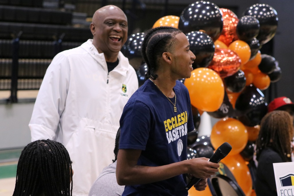 A student in a navy Ecclesia Basketball shirt speaks into a microphone while another person in a white Comets jacket stands behind them. Black and orange balloons decorate the gym.