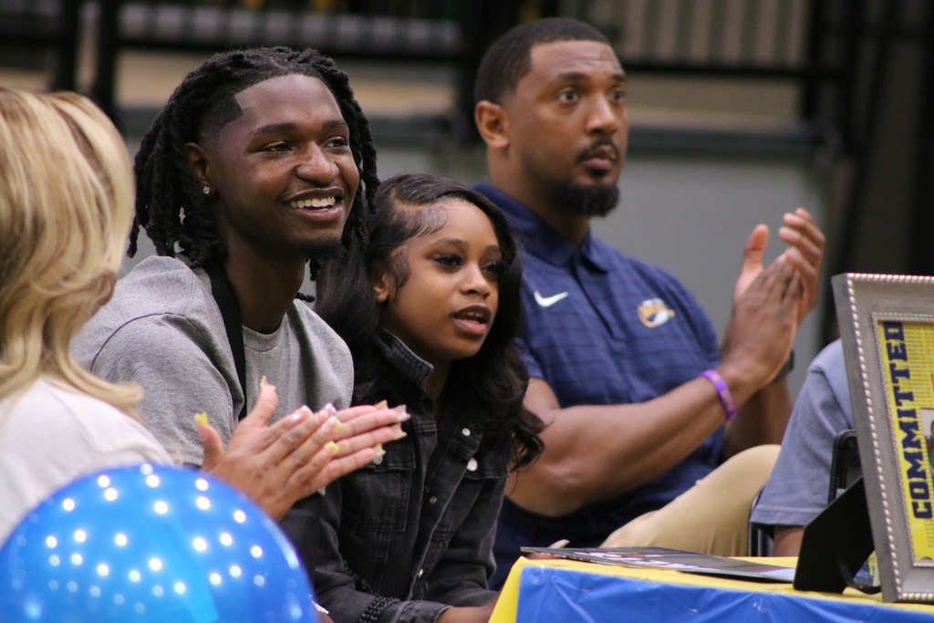 Four people sit at a table during a signing‑day event, clapping and smiling. Blue balloons and a framed “Committed” display are on the table in front of them.