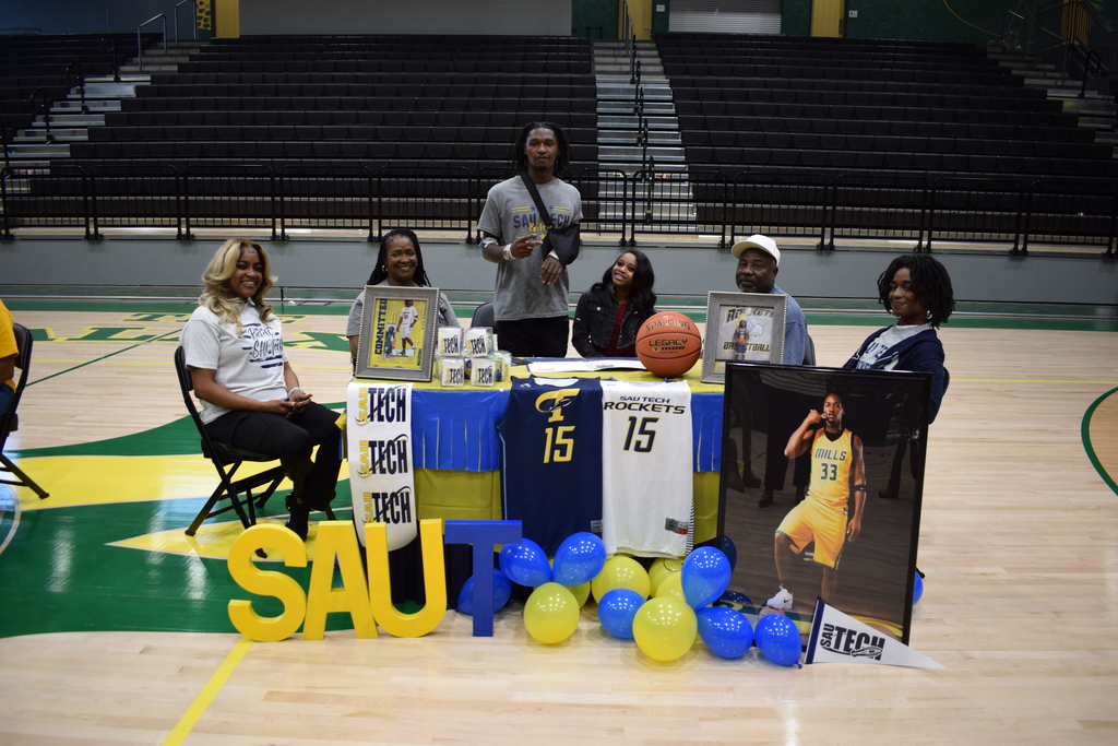 Six people gather around a table covered in blue and yellow SAU Tech decorations, including jerseys, balloons, framed photos, and a basketball, during a signing‑day ceremony.