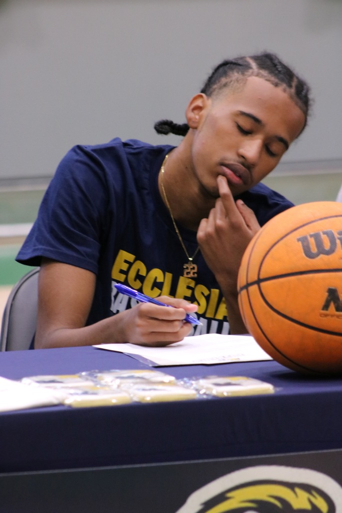 A student in a navy Ecclesia Basketball shirt signs a document at a table with a basketball and a blue tablecloth featuring an eagle logo.