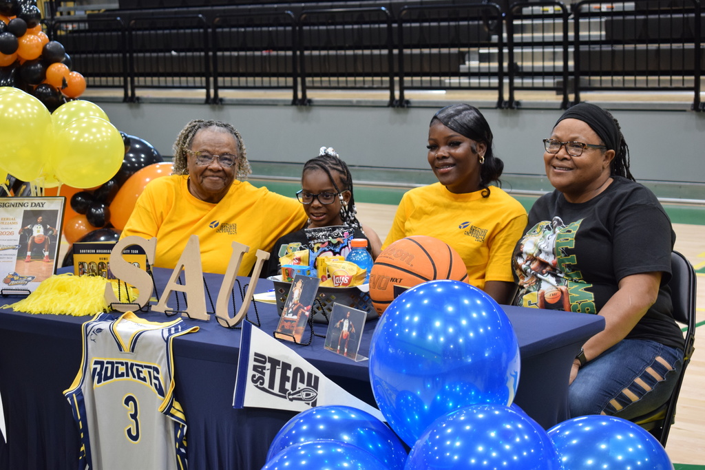 Four people sit at a decorated table with SAU Tech items, balloons, a basketball, and framed photos for a signing‑day celebration. A Rockets jersey hangs from the front of the table.