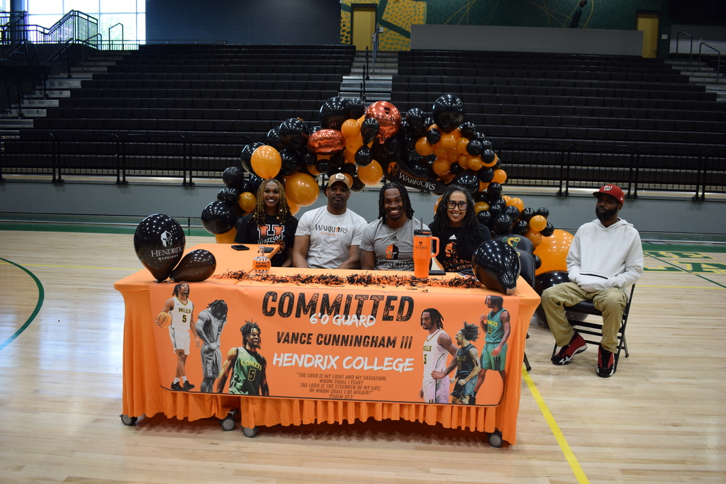 Five people sit behind a table with an orange tablecloth featuring photos and text announcing Vance Cunningham III’s commitment to Hendrix College. A black‑and‑orange balloon arch stands behind them.
