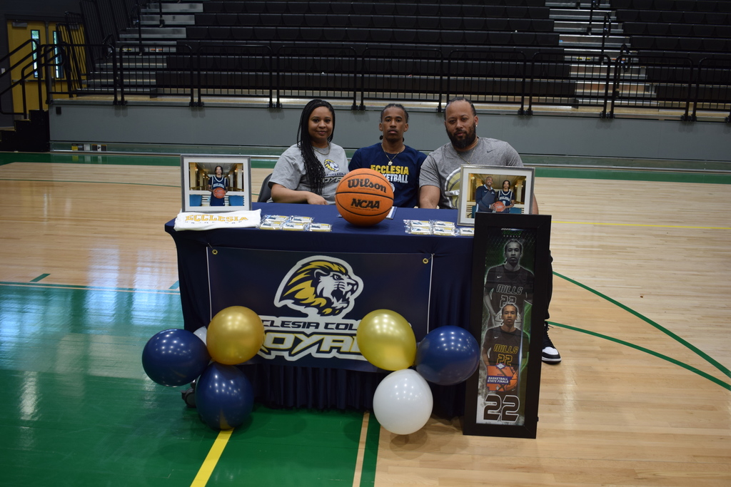 Thre people sit behind a table decorated with Ecclesia College  items, including balloons, framed photos, a basketball, and a jersey. The gym bleachers are visible behind them.