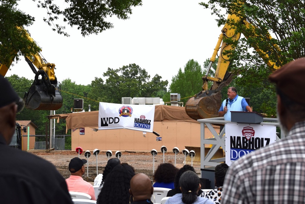 A construction site with cranes holding a banner. A man is speaking at a podium.