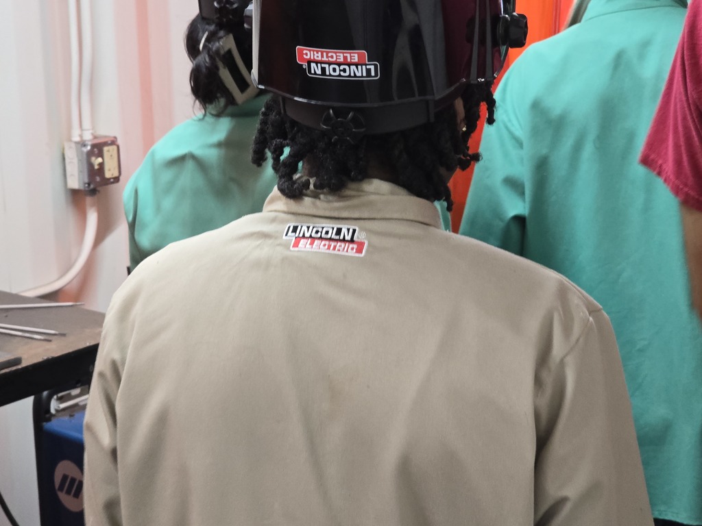 “Student wearing a tan Lincoln Electric welding jacket and helmet stands in a welding booth. Other students in green protective jackets work in the background.”