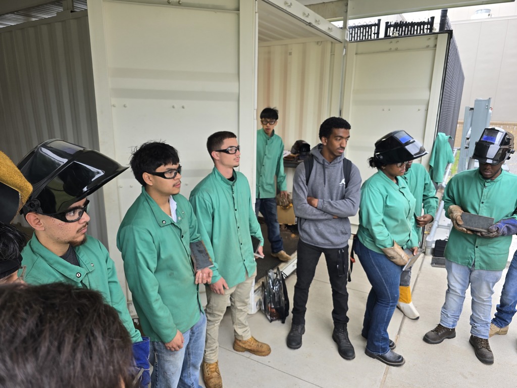 Students in green welding jackets gather around a rectangular metal plate held by one student, examining the weld during an outdoor lesson.