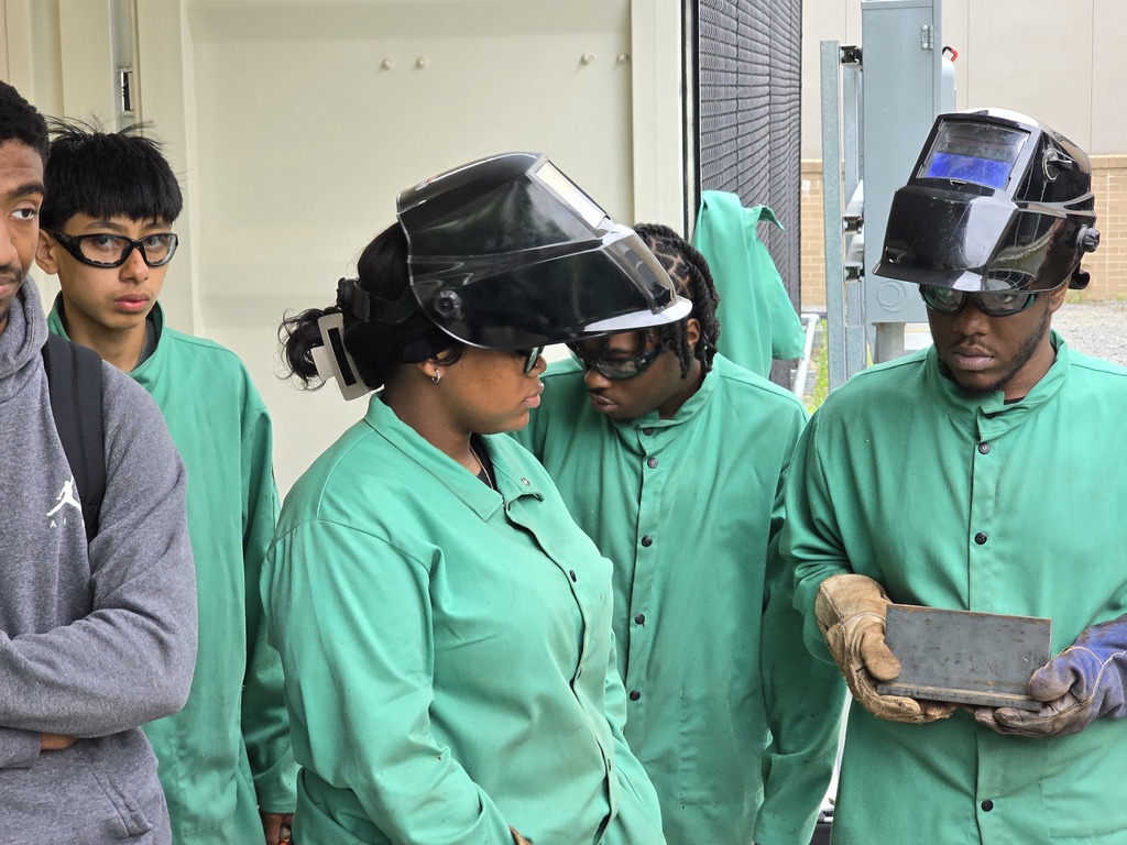 Students in welding jackets gather outdoors around a rectangular metal plate held by one student, examining the weld during an outdoor lesson.