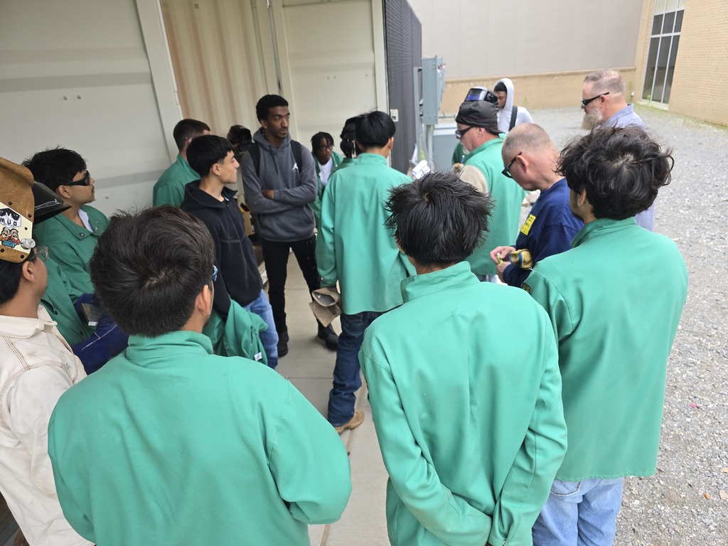 Students in welding gear stand near a building with large windows. One student holds a welded metal piece while others observe. Two visitors with yellow badges stand nearby.