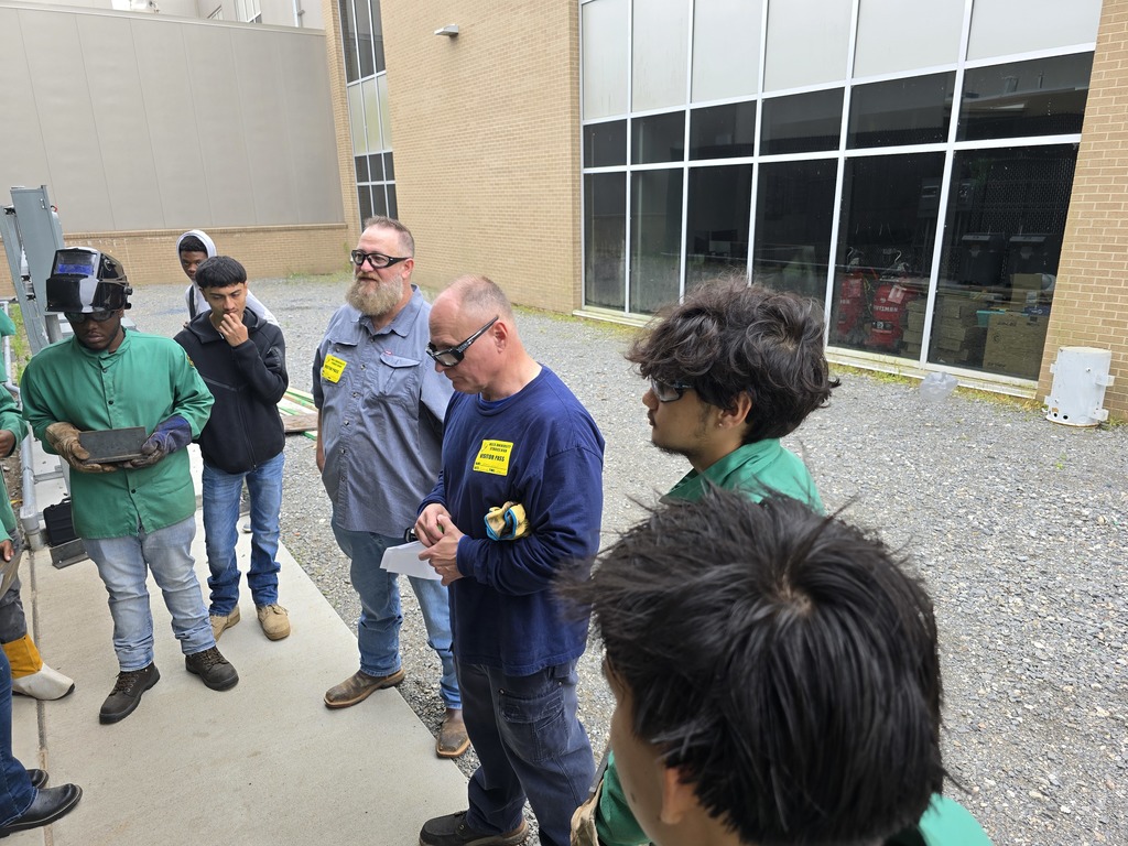 Group of students in green welding jackets and safety glasses stand outside near a workshop area. One student holds a metal piece while the group listens to an instructor.