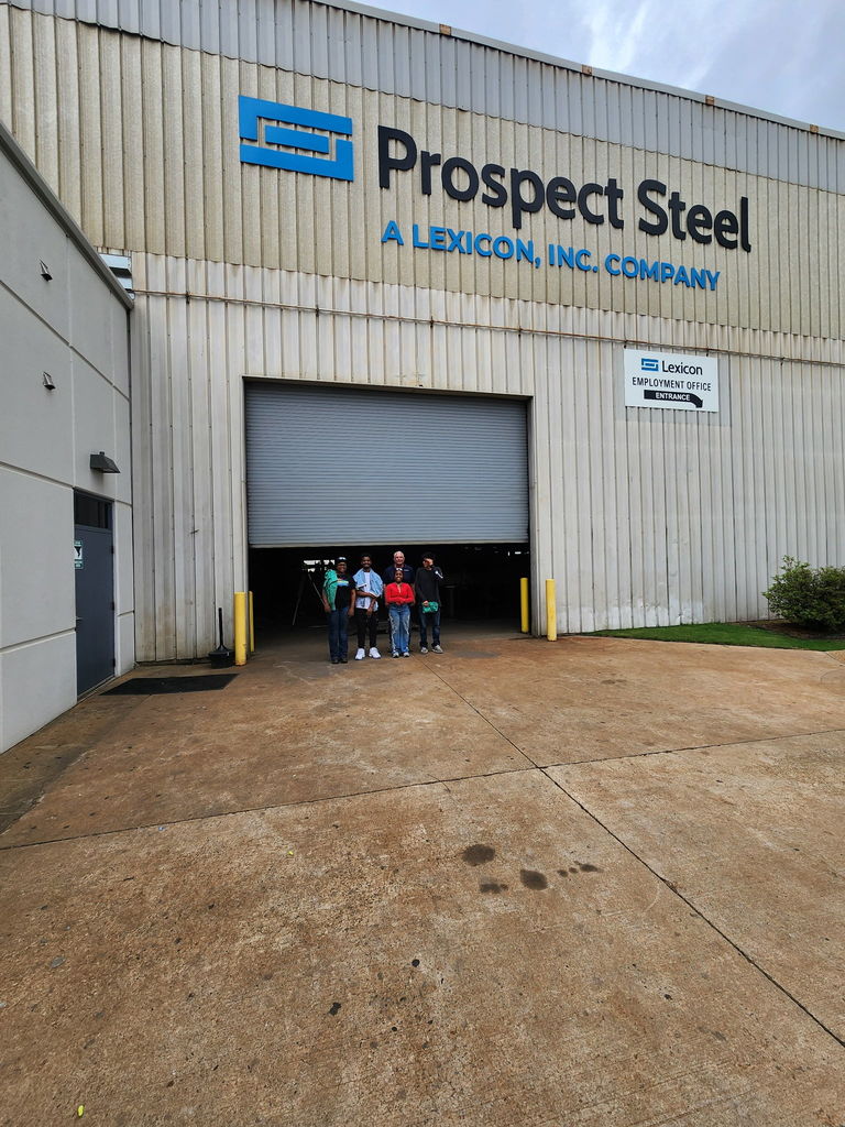 Exterior of a large metal building with signage reading ‘Prospect Steel — A Lexicon, Inc. Company.’ A smaller sign marks the employment office entrance. Several people stand on the concrete area in front of the building.