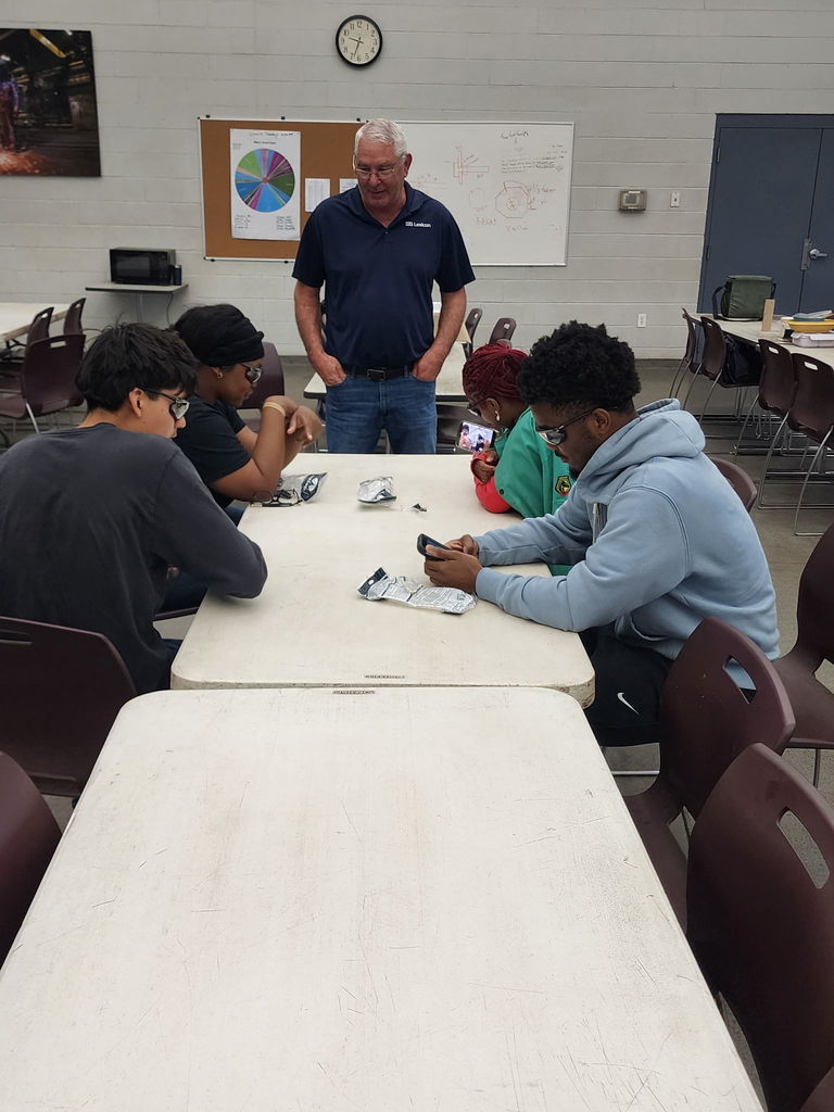 Four students sit at a table working with small electronic components while an instructor in a navy Lincoln shirt stands nearby. A bulletin board, clock, and microwave are visible in the background.