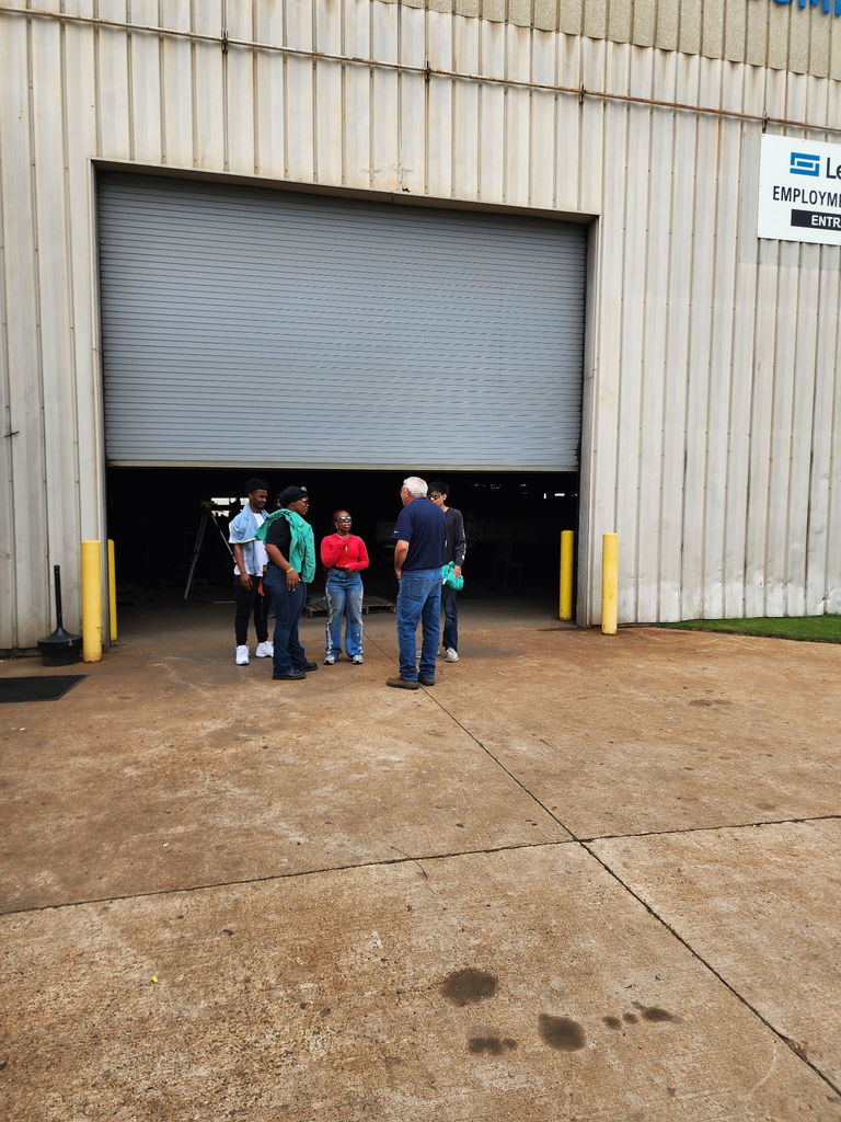 Five people stand talking outside a large metal industrial building with a partially open roll‑up door. A sign reading ‘Employment Entrance’ are visible near the doorway