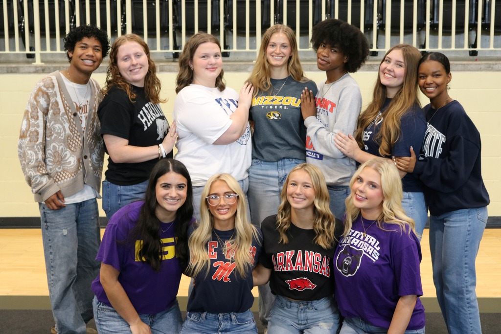Eleven students pose in two rows wearing shirts from various colleges inside a gym‑like space.