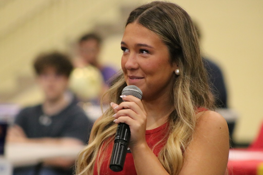 A person in a red sleeveless top speaks into a microphone at an indoor event with people seated behind them.