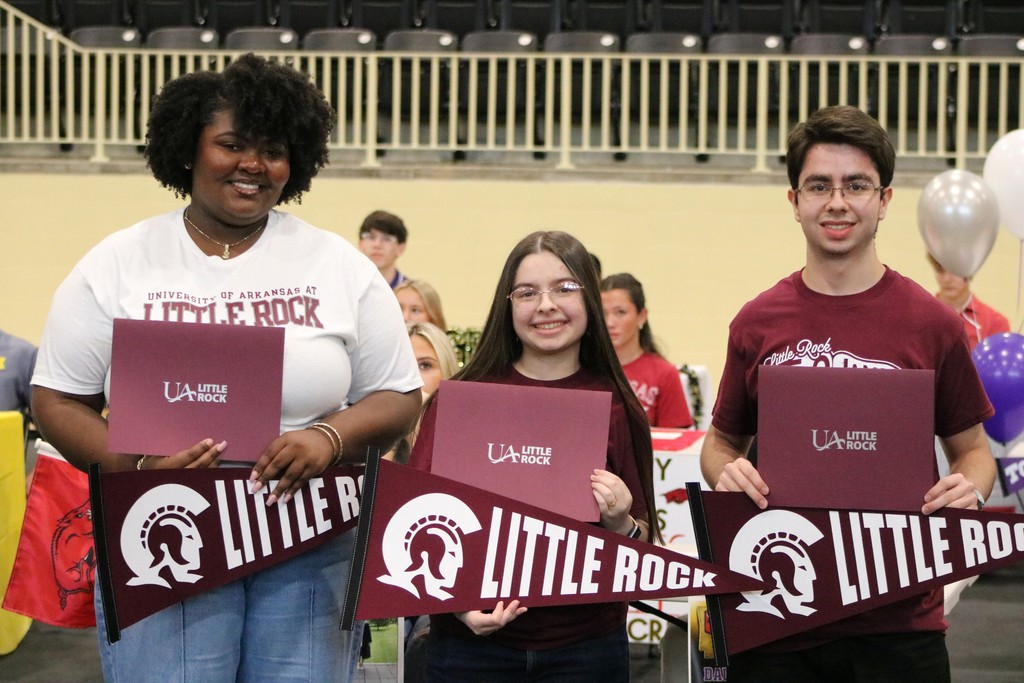 Three individuals hold UA Little Rock pennants and folders while standing in front of balloons and seated attendees.