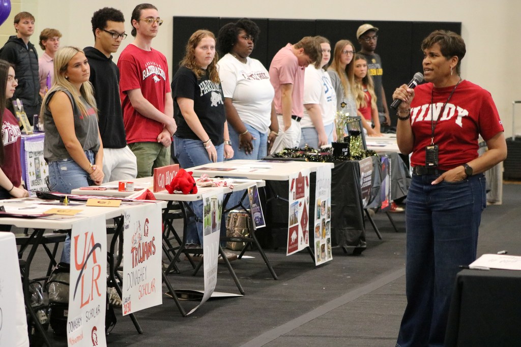 A group stands behind decorated tables displaying college and scholarship materials while a person in a red shirt speaks into a microphone.