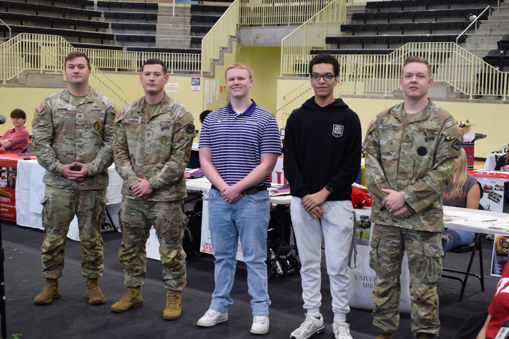 Three people in Army uniforms and two students stand in a row inside a gym during a military or informational event.