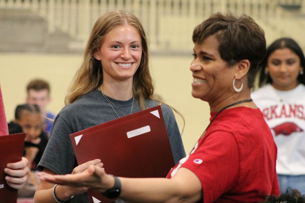 Two individuals talk while one holds a maroon folder; several people and college‑themed shirts are visible in the background.