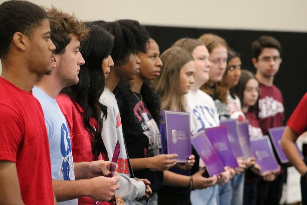 A line of students holds purple folders during an indoor recognition ceremony.
