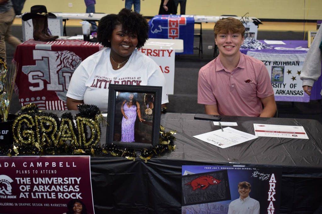 Two students sit at decorated tables during a signing event, each with posters and items showing their chosen universities.