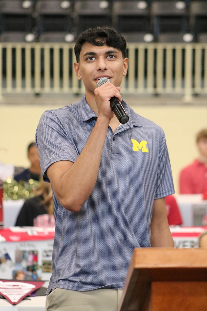 A student wearing a blue University of Michigan shirt speaks into a microphone near a wooden podium at an indoor event.