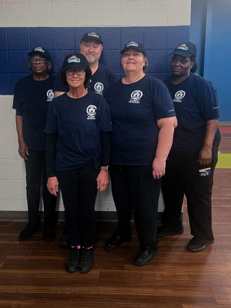 Five cafeteria staff members wearing matching navy Student Nutrition shirts and “Champion” caps stand against a tiled wall.