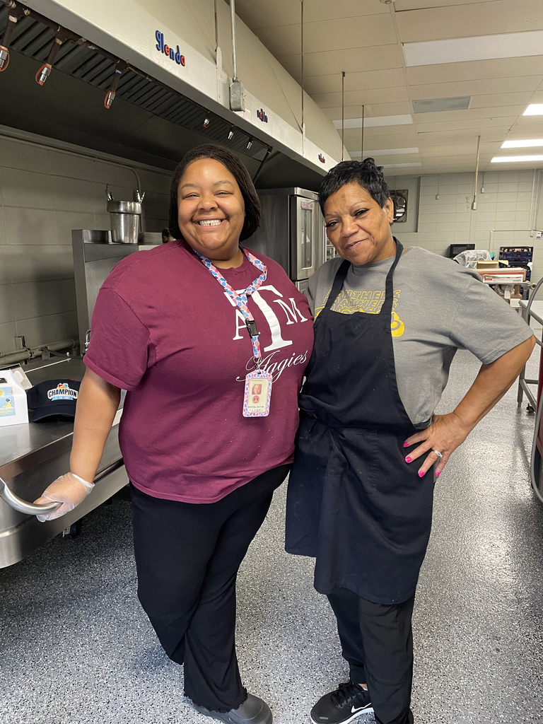 Two cafeteria staff members stand side by side in a stainless‑steel commercial kitchen with ovens and equipment behind them.
