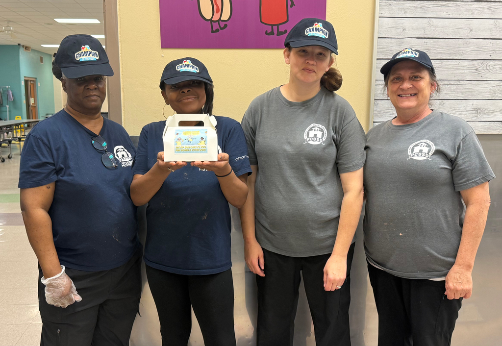 Four cafeteria staff members wearing “Champion” caps pose together; one holds a small box with a School Lunch Hero message.